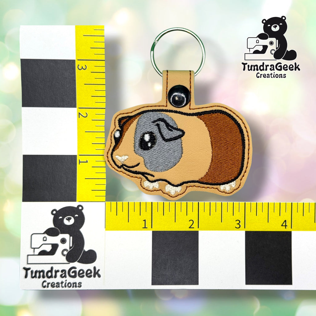 Guinea pig keychain shown against a black-and-white checkered background with a yellow ruler for scale, measuring approximately 3.75 inches wide by 3.25 inches tall.  ---
