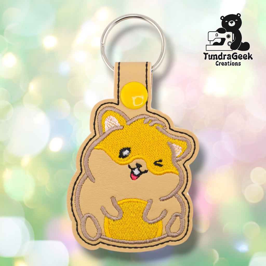 Tan faux leather keychain shaped like a chubby sitting hamster. Golden yellow embroidery covers the body; the face features one open eye, one winking eye, a small pink tongue, and white inner ear detail. Yellow snap closure and silver key ring at top. TundraGeek Creations logo in upper right corner.