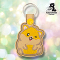 Tan faux leather keychain shaped like a chubby sitting hamster. Golden yellow embroidery covers the body; the face features one open eye, one winking eye, a small pink tongue, and white inner ear detail. Yellow snap closure and silver key ring at top. TundraGeek Creations logo in upper right corner.