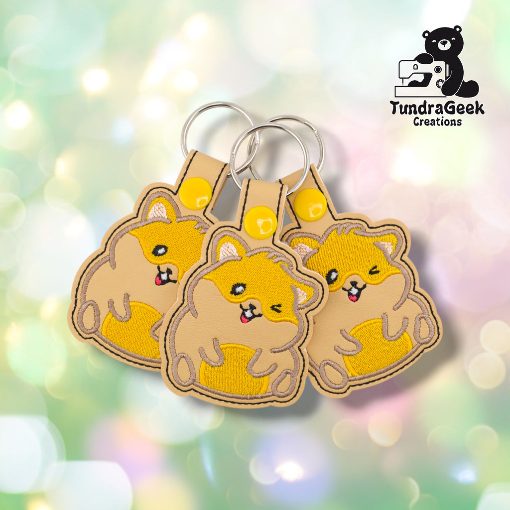 Three hamster keychains fanned out on a pastel bokeh background, showing slightly varied angles of the same design. All three feature matching golden yellow embroidery, tan base, and yellow snap closures.