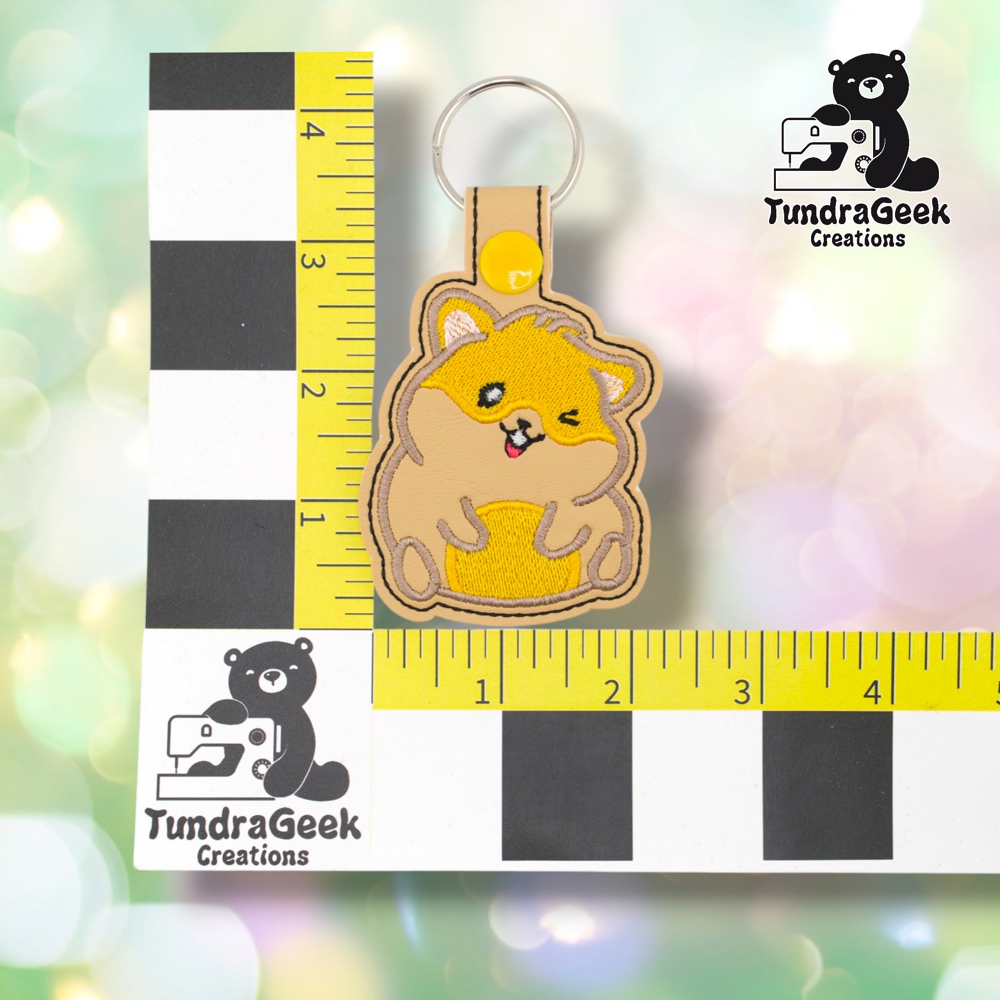 Hamster keychain positioned against a TundraGeek Creations branded ruler card showing yellow tape measure. Keychain body measures approximately 2.25 inches wide by 3.5 inches tall, not including hardware.