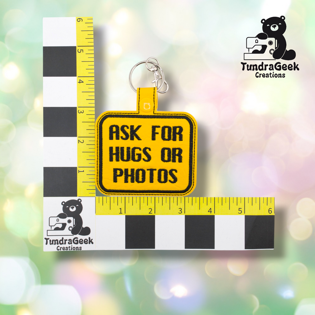 Yellow embroidered vinyl keychain badge reading “ASK FOR HUGS OR PHOTOS” in black text with a black border. Shown against a ruler indicating approximately 3 inches wide and 2.5 inches tall. Silver lobster claw clip and yellow snap hardware at top