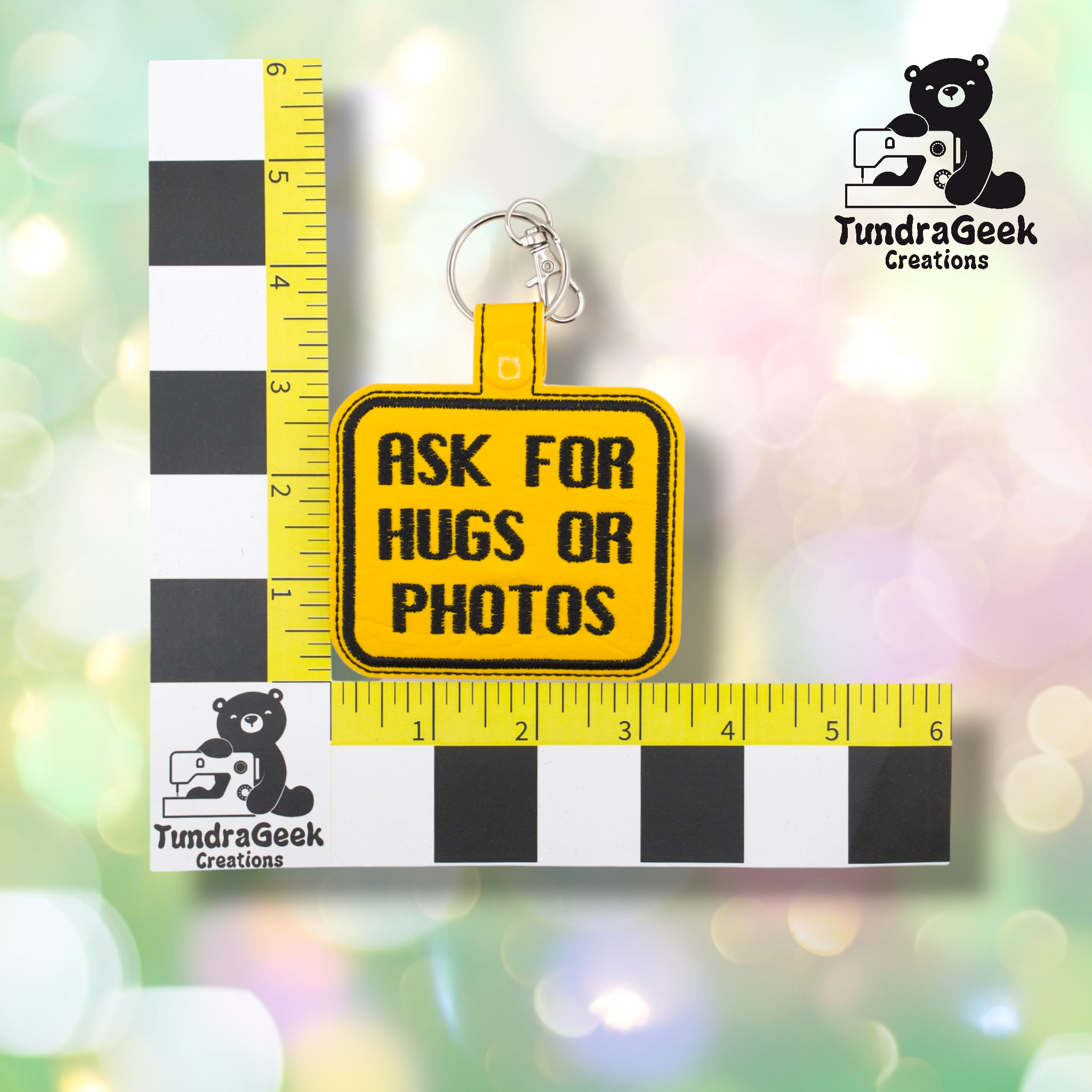 Yellow embroidered vinyl keychain badge reading “ASK FOR HUGS OR PHOTOS” in black text with a black border. Shown against a ruler indicating approximately 3 inches wide and 2.5 inches tall. Silver lobster claw clip and yellow snap hardware at top