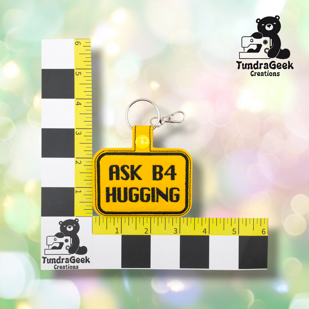 Yellow embroidered vinyl keychain badge reading “ASK B4 HUGGING” in black text with a black border, shown against a ruler indicating approximately 3 inches wide and 2 inches tall. Silver lobster claw clip and ring hardware attached at top.