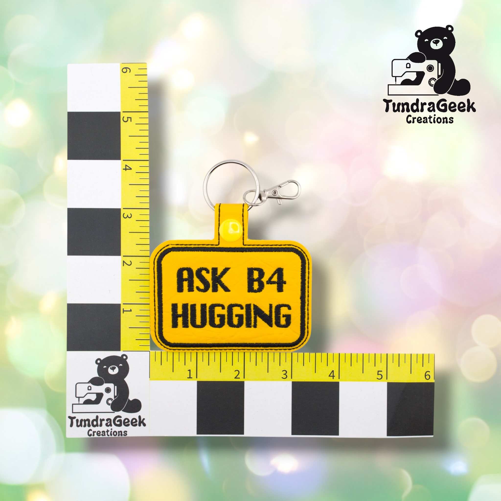 Yellow embroidered vinyl keychain badge reading “ASK B4 HUGGING” in black text with a black border, shown against a ruler indicating approximately 3 inches wide and 2 inches tall. Silver lobster claw clip and ring hardware attached at top.