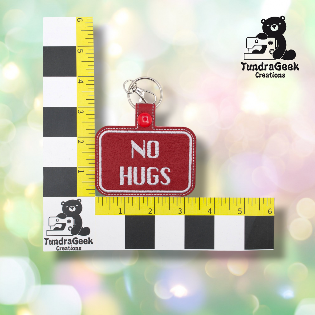 Dark red embroidered vinyl keychain badge reading “NO HUGS” in white text with a white border. Shown against a ruler indicating approximately 3 inches wide and 2 inches tall. Silver lobster claw clip and red snap hardware at top