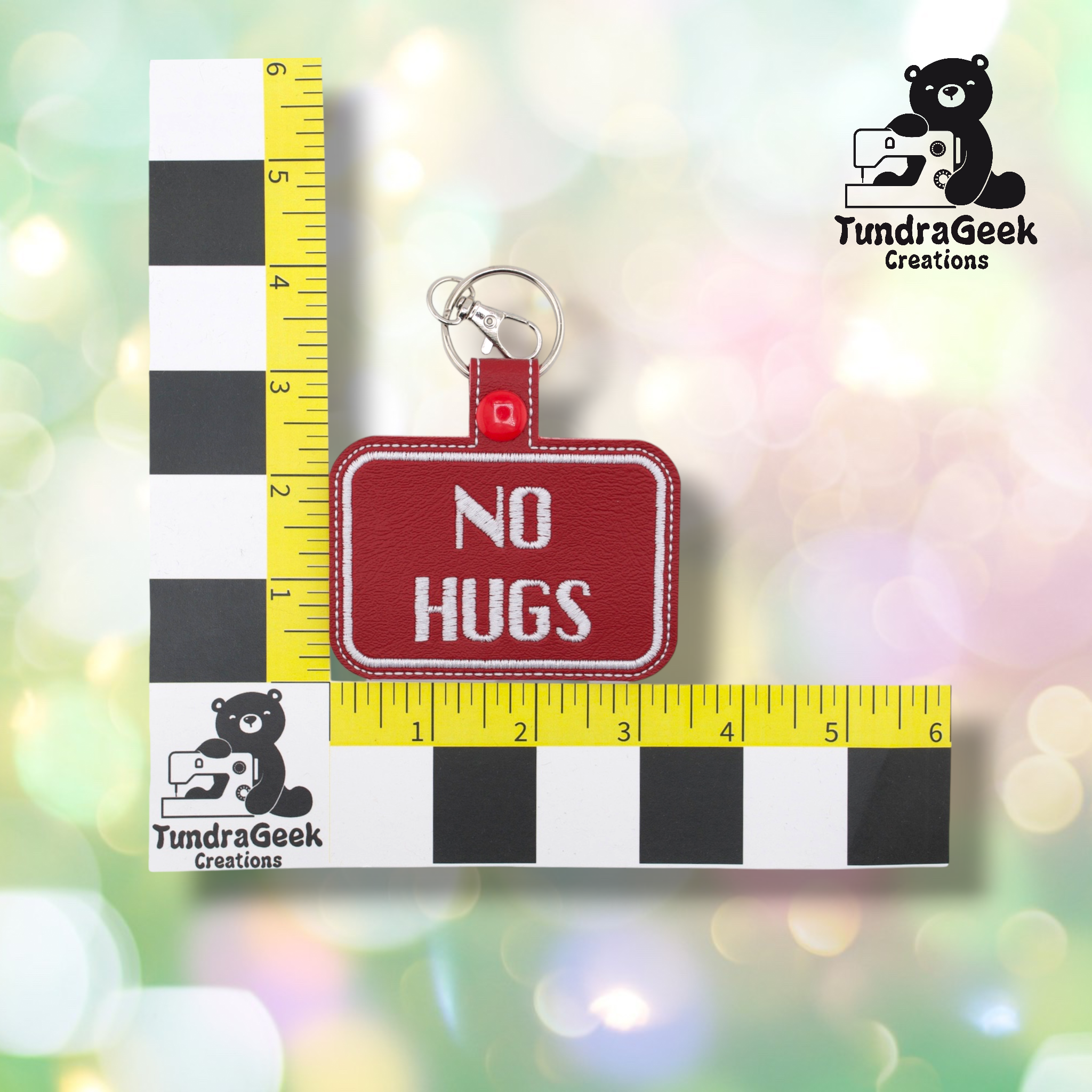 Dark red embroidered vinyl keychain badge reading “NO HUGS” in white text with a white border. Shown against a ruler indicating approximately 3 inches wide and 2 inches tall. Silver lobster claw clip and red snap hardware at top