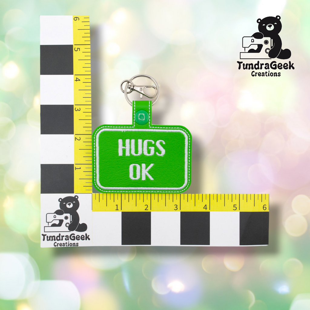Bright green embroidered vinyl keychain badge reading “HUGS OK” in white text with a white border. Shown against a ruler indicating approximately 3 inches wide and 2 inches tall. Silver lobster claw clip and green snap hardware at top