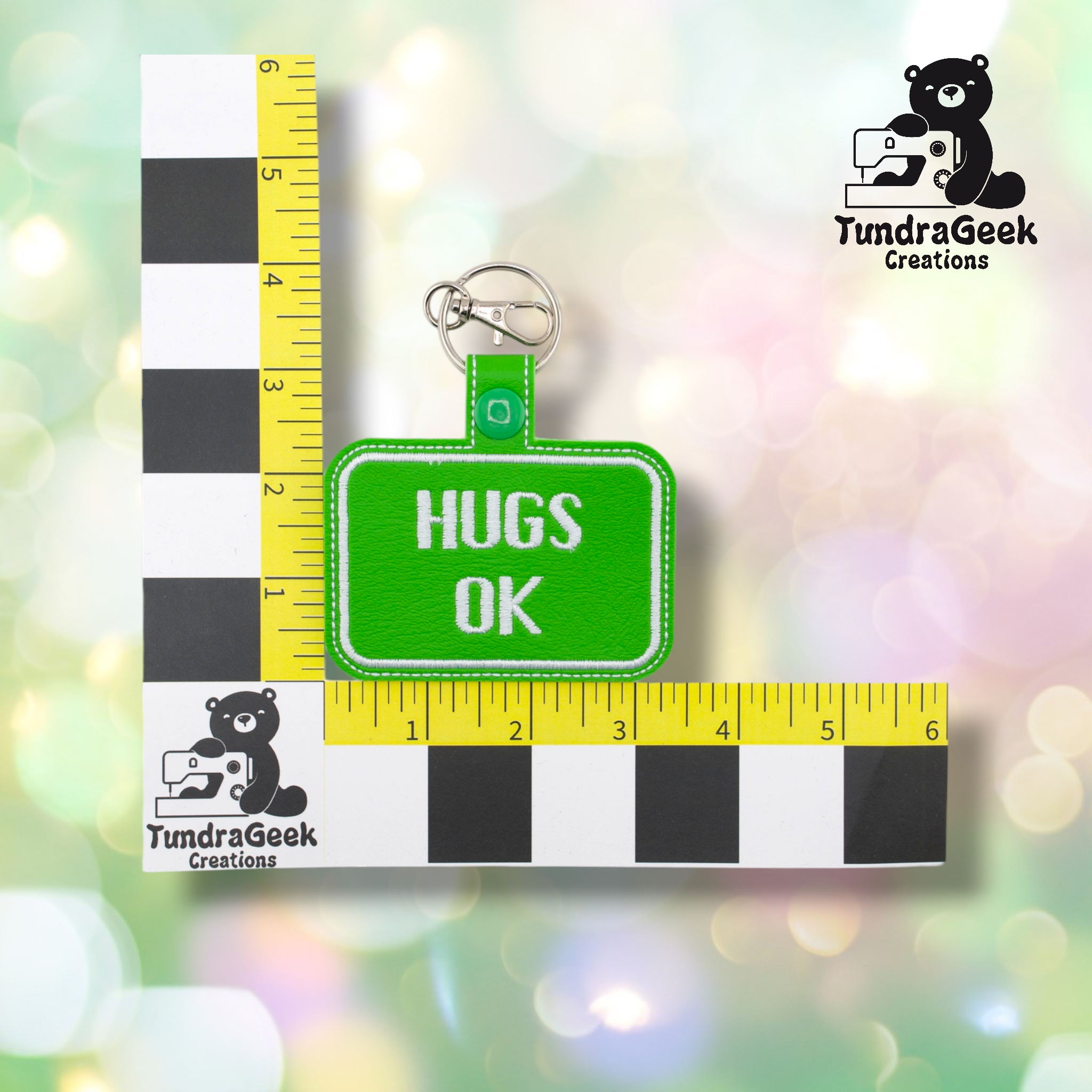 Bright green embroidered vinyl keychain badge reading “HUGS OK” in white text with a white border. Shown against a ruler indicating approximately 3 inches wide and 2 inches tall. Silver lobster claw clip and green snap hardware at top