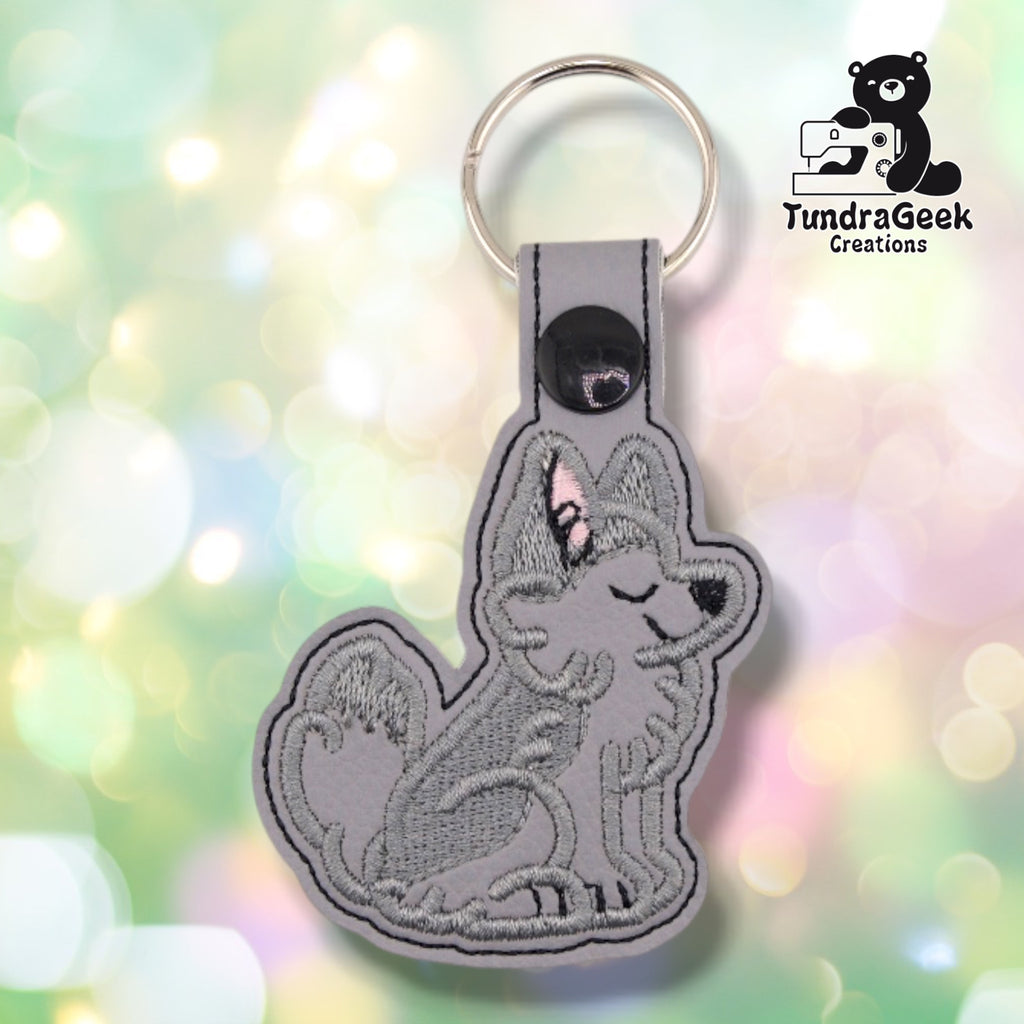 Light grey faux leather keychain cut to the shape of a sitting husky, embroidered in darker grey thread. The dog faces right with its eyes closed and its muzzle tipped upward, one ear lined in pink, a black nose, and a thick curled tail behind it. A black plastic snap closes the tab around a silver split ring.