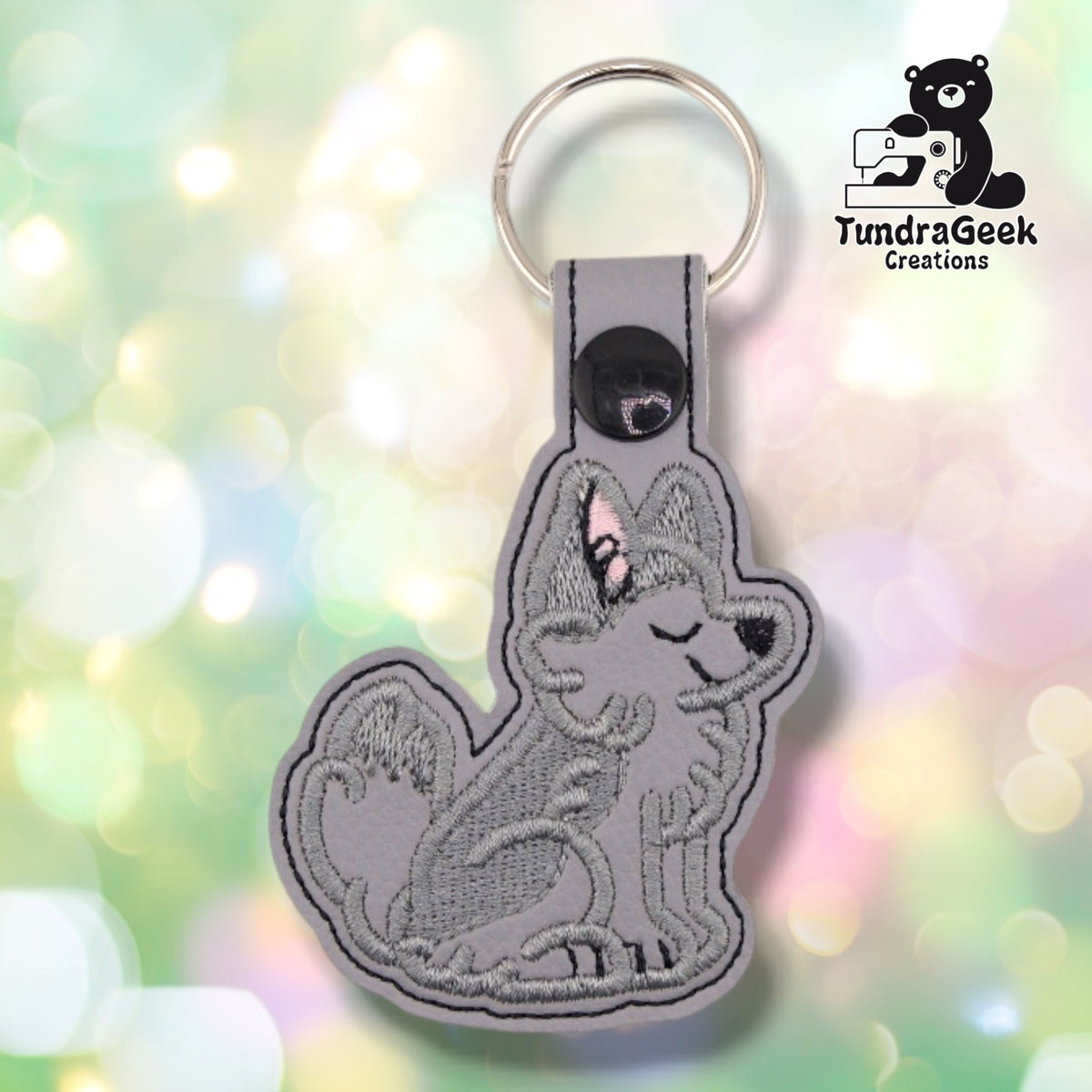 Light grey faux leather keychain cut to the shape of a sitting husky, embroidered in darker grey thread. The dog faces right with its eyes closed and its muzzle tipped upward, one ear lined in pink, a black nose, and a thick curled tail behind it. A black plastic snap closes the tab around a silver split ring.