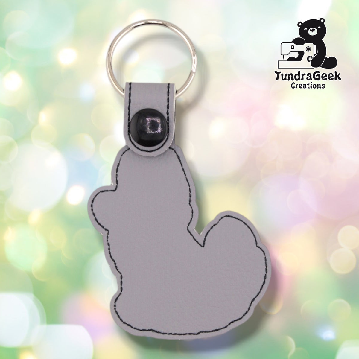 The back of the husky keychain, plain light grey faux leather with a black stitched outline following the husky silhouette. The snap tab, black plastic snap and silver split ring are at the top.