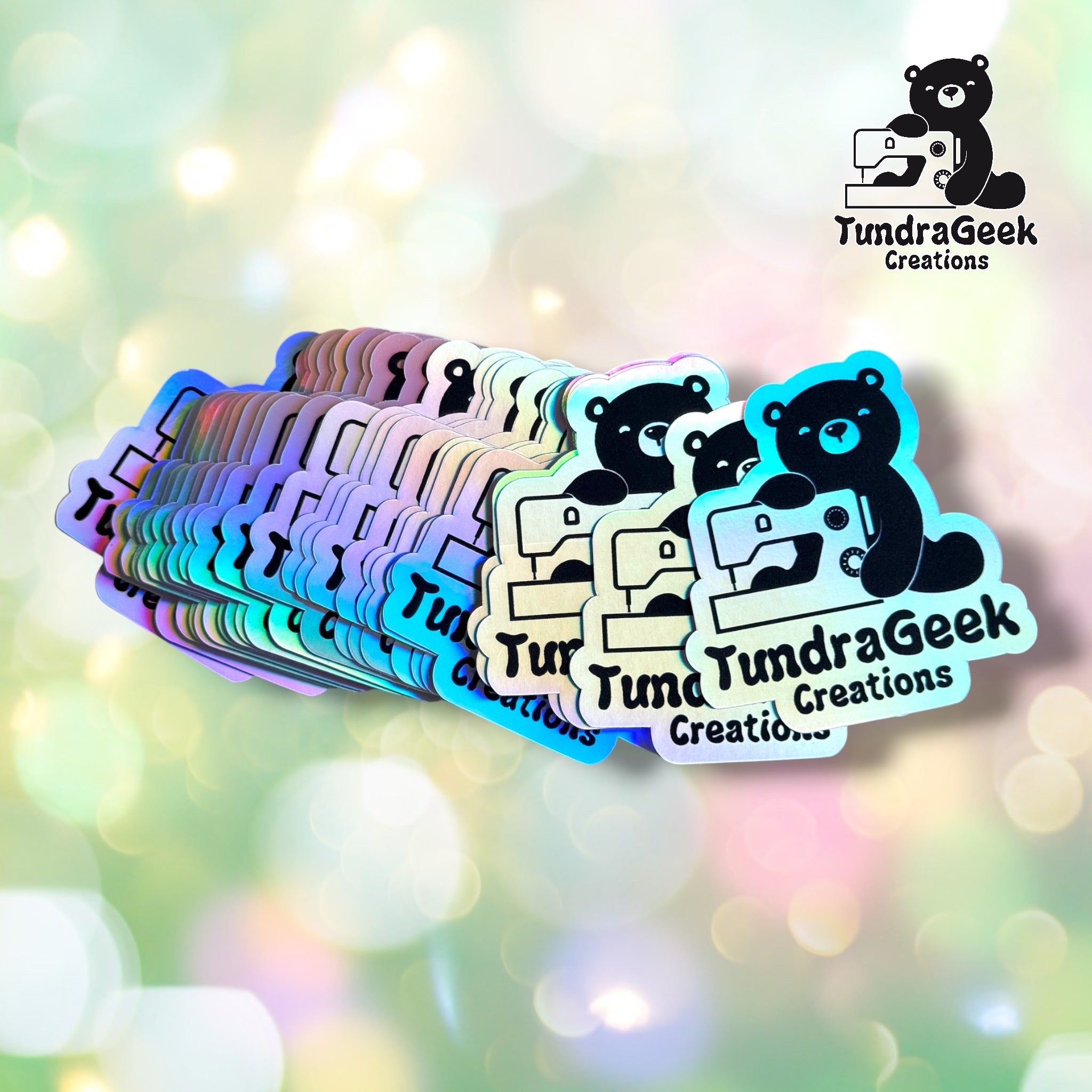 A fan of small die-cut stickers featuring a black cartoon bear holding a sewing machine, with the text 'TundraGeek Creations' on the stickers. The stickers have holographic, iridescent edges and are displayed against a soft, colorful bokeh background.