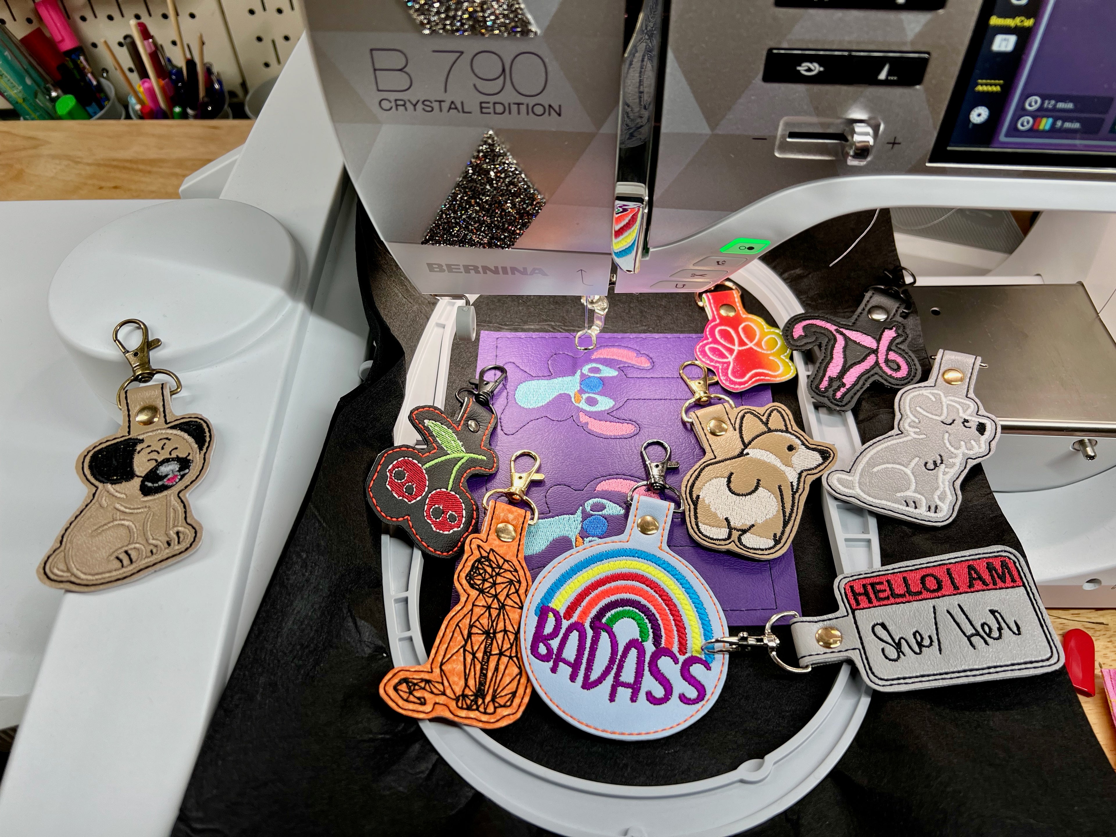 Embroidered keychains displayed on a sewing machine hoop, including a rainbow "BADASS" circle, animals, and a "HELLO I AM she/her" name tag on purple fabric.