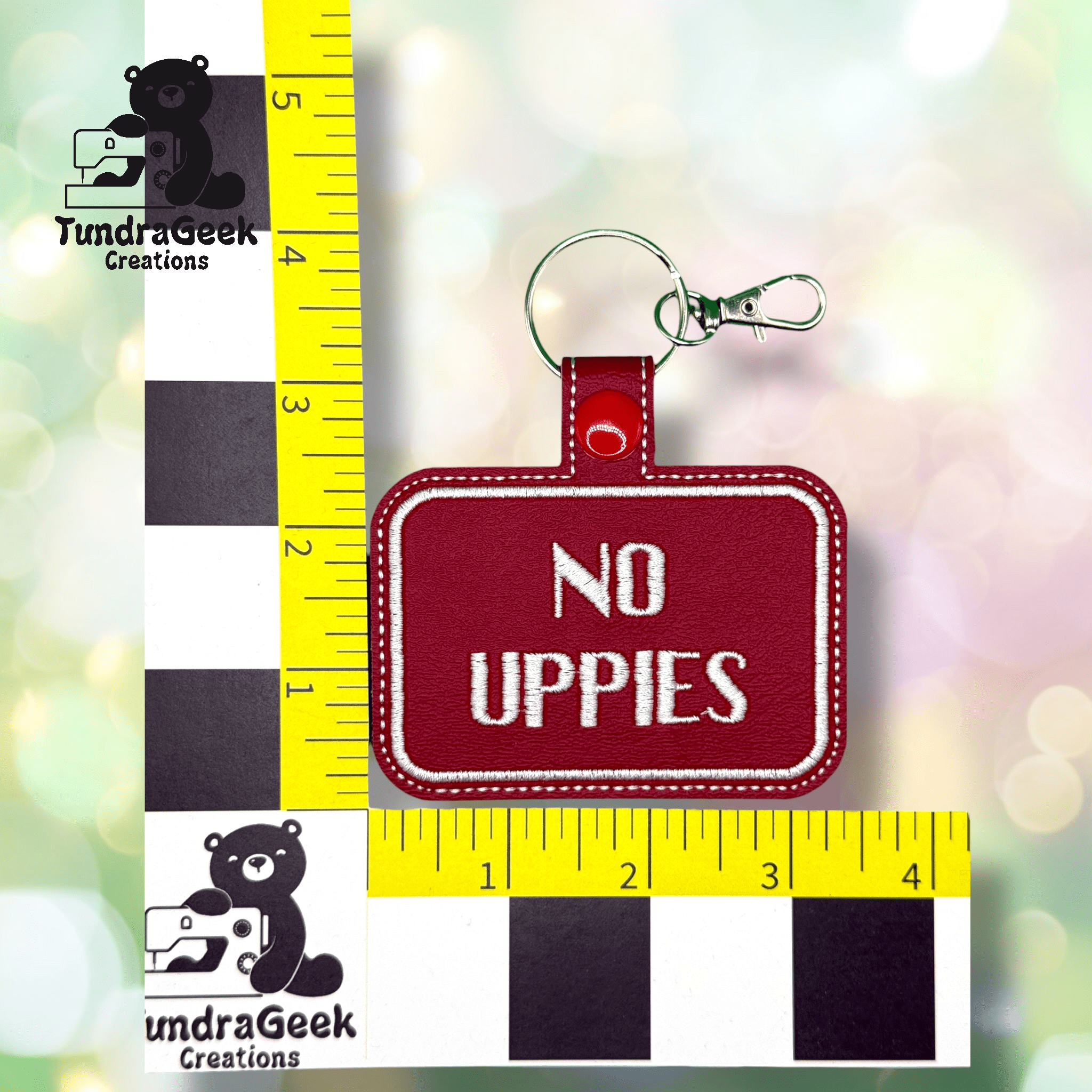 Red keychain with white stitched text reading NO UPPIES, placed against a yellow and black measurement grid showing a height of about 3 inches.