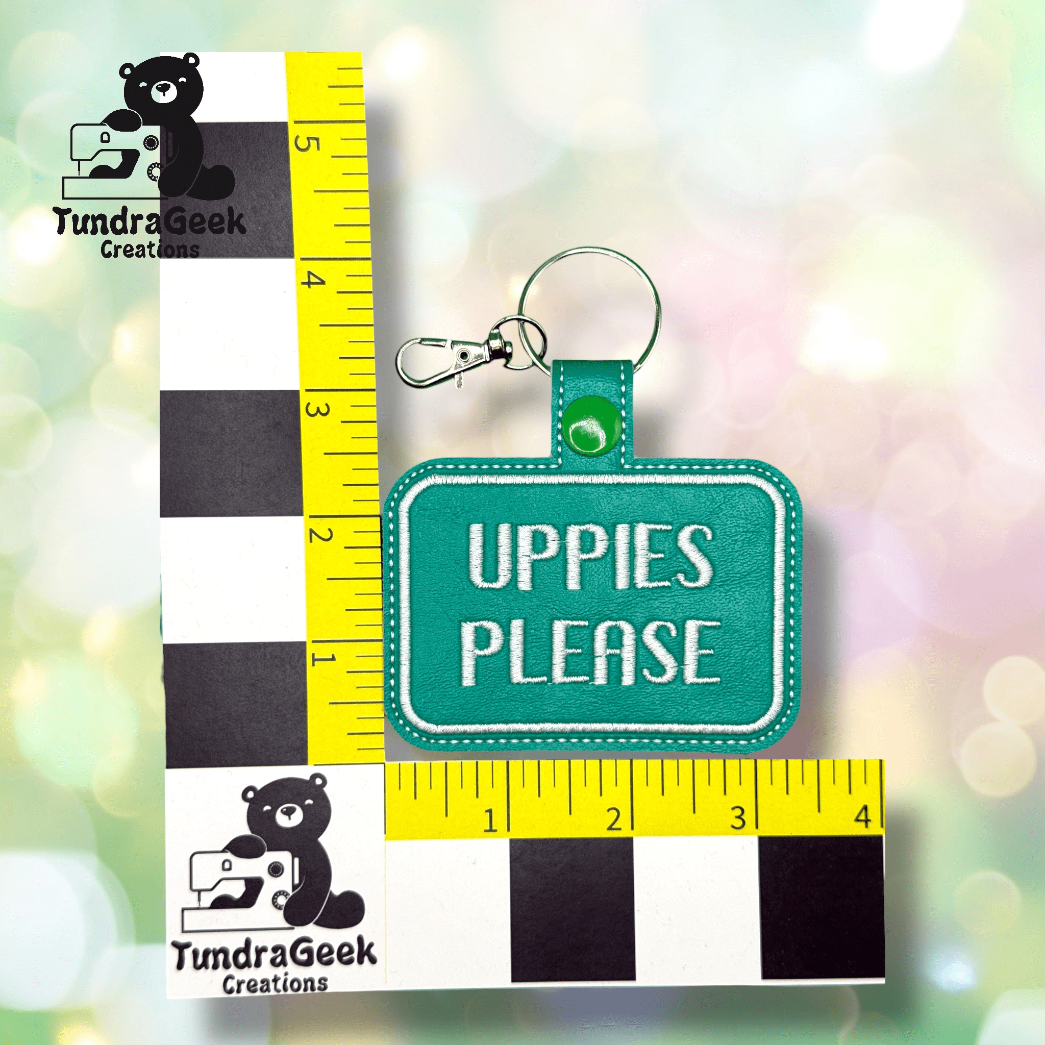 A green keychain tag with white text reading UPPIES PLEASE is shown against a checkered ruler for size reference.