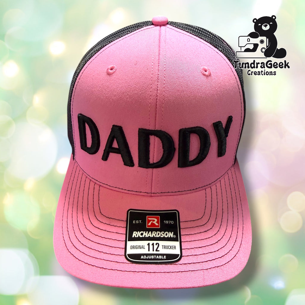Pink Richardson Original 112 trucker cap with black 3D embroidered "DADDY" lettering and charcoal mesh back panel; soft blurred green and white backdrop.
