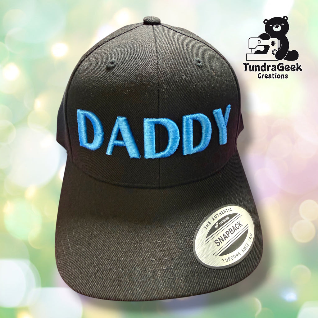Black snapback cap with blue 3D embroidered "DADDY" lettering; soft-focus bokeh background with TundraGeek Creations logo in top right corner.