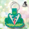 Teal faux leather keychain shaped like a large carnivorous plant with its mouth open wide. White embroidery forms the serrated teeth; the interior is embroidered in pink/coral. Green stem embroidery curves up both sides and light green leaf embroidery spreads along the base. Green plastic snap closure and silver key ring at top. TundraGeek Creations logo in upper right corner.