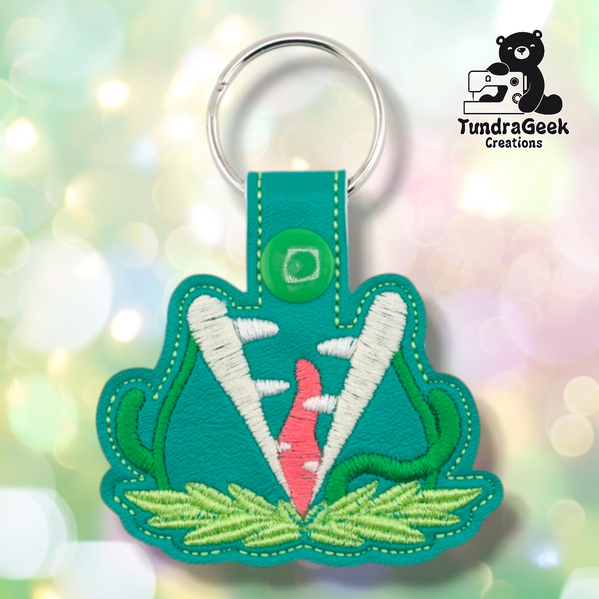Teal faux leather keychain shaped like a large carnivorous plant with its mouth open wide. White embroidery forms the serrated teeth; the interior is embroidered in pink/coral. Green stem embroidery curves up both sides and light green leaf embroidery spreads along the base. Green plastic snap closure and silver key ring at top. TundraGeek Creations logo in upper right corner.