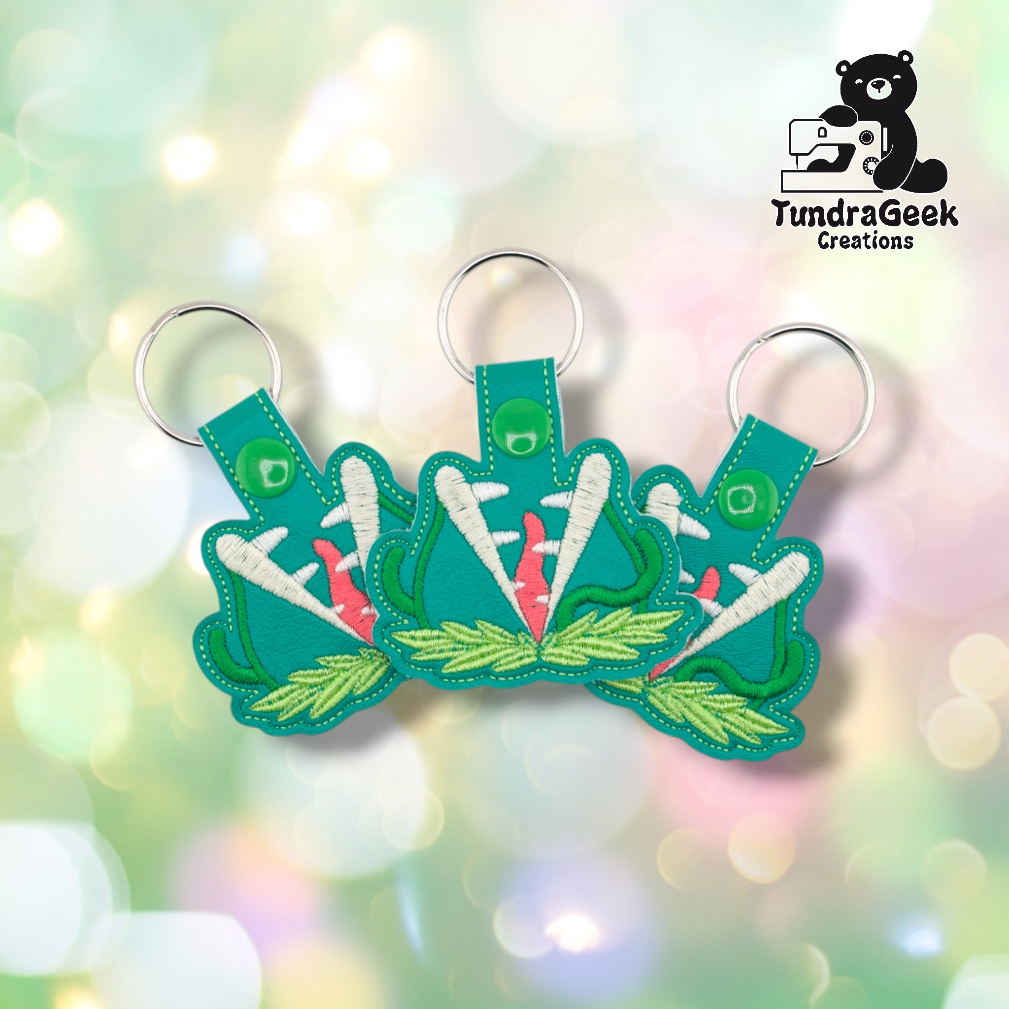 Three killer plant keychains arranged together on a pastel bokeh background, showing the design from slightly varied angles. All three feature matching teal faux leather, white tooth embroidery, and green snap closures.