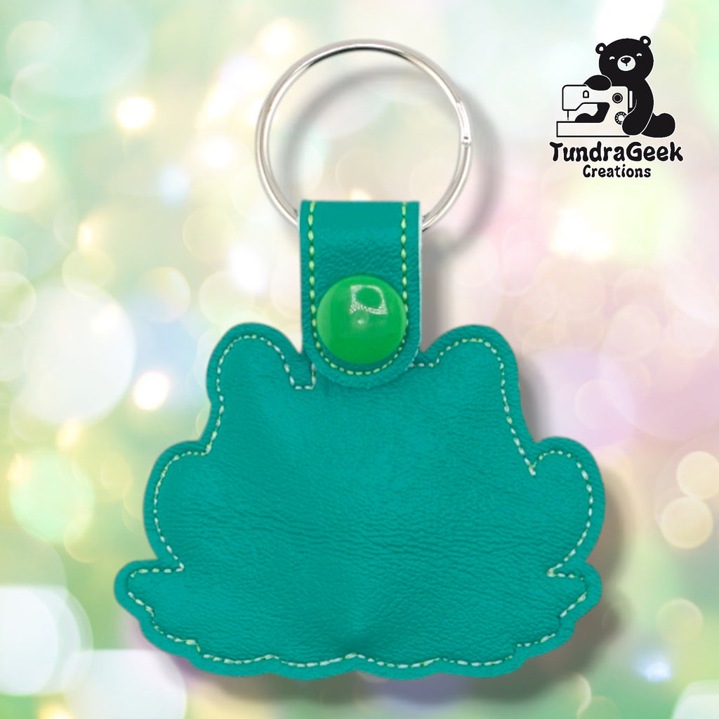 Plain back of the killer plant keychain showing teal faux leather and yellow-green outline stitching following the plant silhouette. Green plastic snap closure at top. No embroidery on reverse.