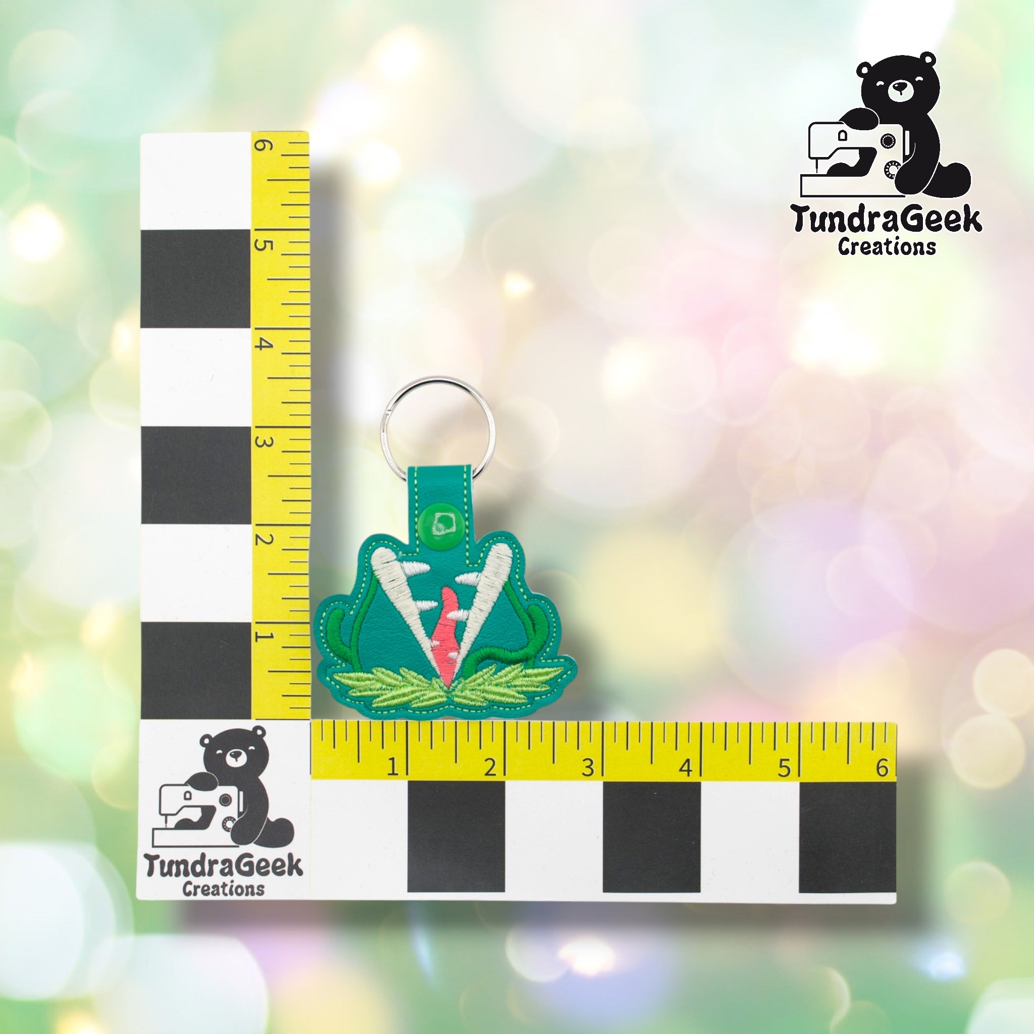 Killer plant keychain positioned against a TundraGeek Creations branded ruler card showing yellow tape measure. Keychain body measures approximately 2.75 inches wide by 2 inches tall, not including hardware.