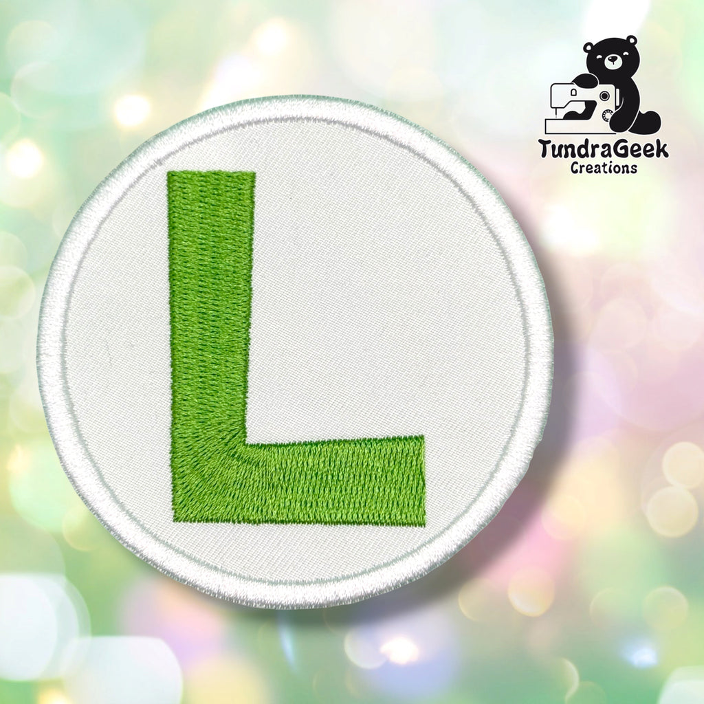 A circular embroidered patch featuring a bright green capital letter 'L' on a white fabric background with a stitched border, shown against a soft pastel bokeh background. The TundraGeek Creations logo is visible in the top right corner.