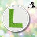 A circular embroidered patch featuring a bright green capital letter 'L' on a white fabric background with a stitched border, shown against a soft pastel bokeh background. The TundraGeek Creations logo is visible in the top right corner.