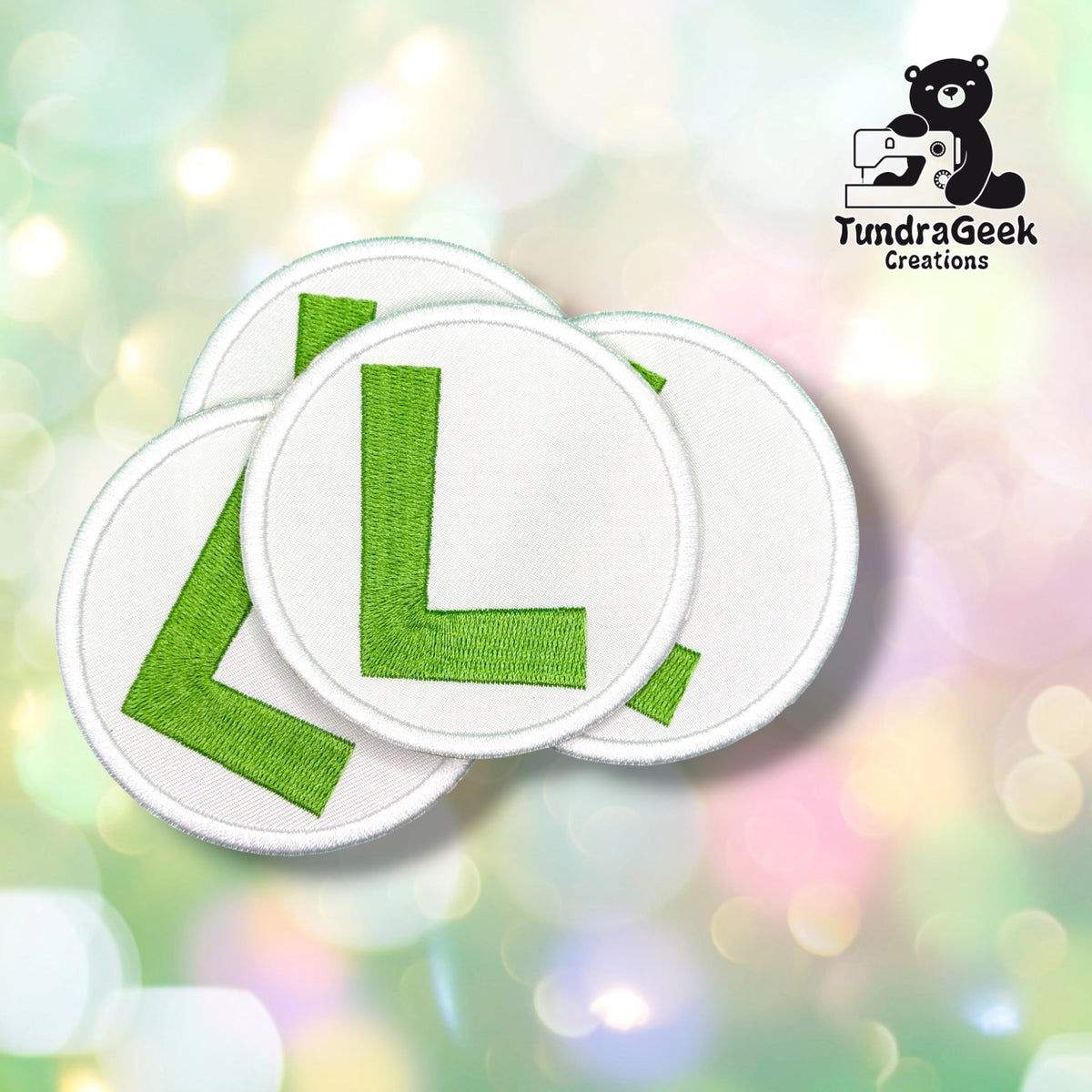 Four circular white embroidered patches with green letter 'L' designs, arranged in a fan shape against a colorful pastel bokeh background. A TundraGeek Creations logo is visible in the top right corner.
