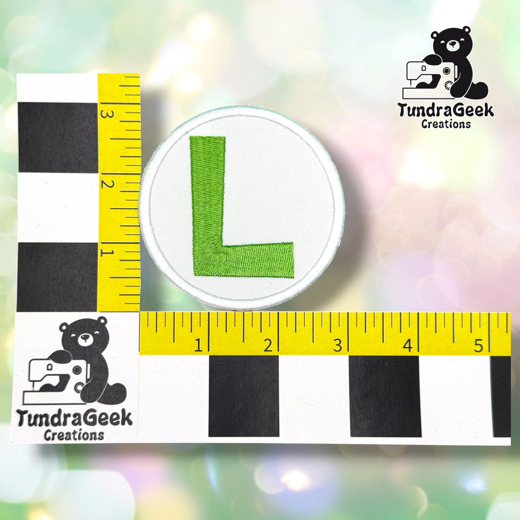 A circular embroidered patch featuring a green letter 'L' on a white background, shown with a yellow measuring tape and black-and-white checkerboard scale card for size reference. The TundraGeek Creations bear logo appears in the top right and bottom left against a soft pastel bokeh background.