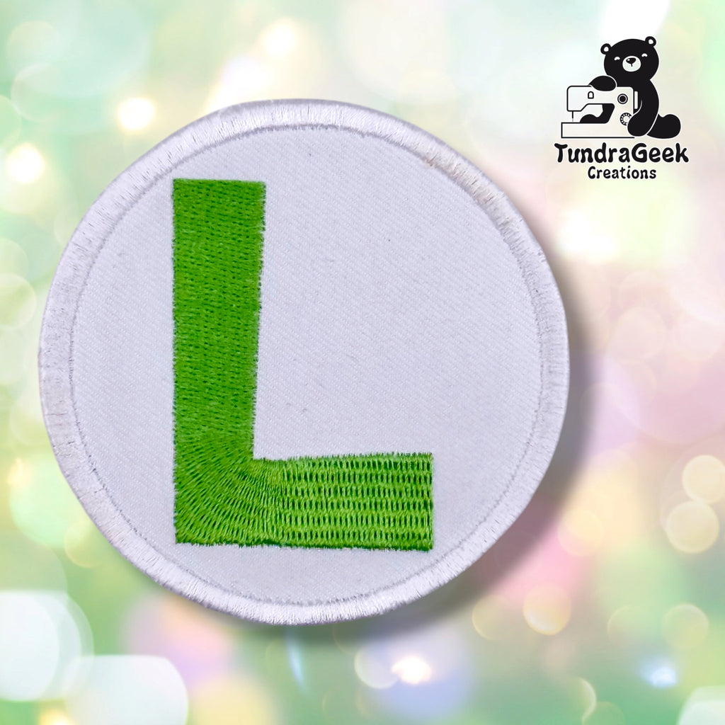A circular embroidered patch featuring a bright green letter 'L' on a white background with a white stitched border, set against a soft pastel bokeh background. The TundraGeek Creations logo is visible in the top right corner.