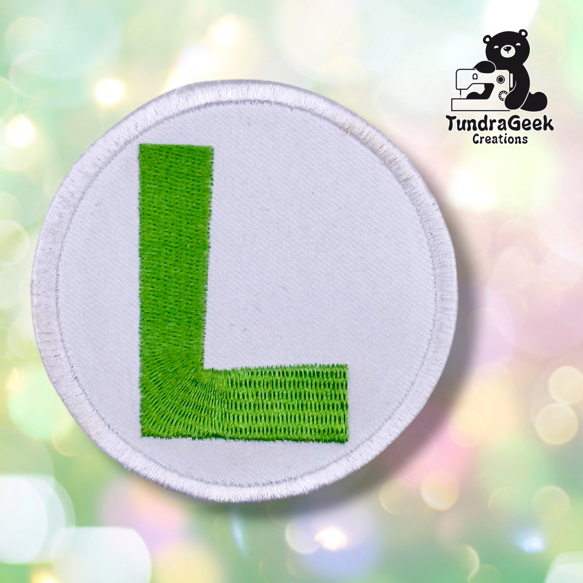 A circular embroidered patch featuring a bright green letter 'L' on a white background with a white stitched border, set against a soft pastel bokeh background. The TundraGeek Creations logo is visible in the top right corner.