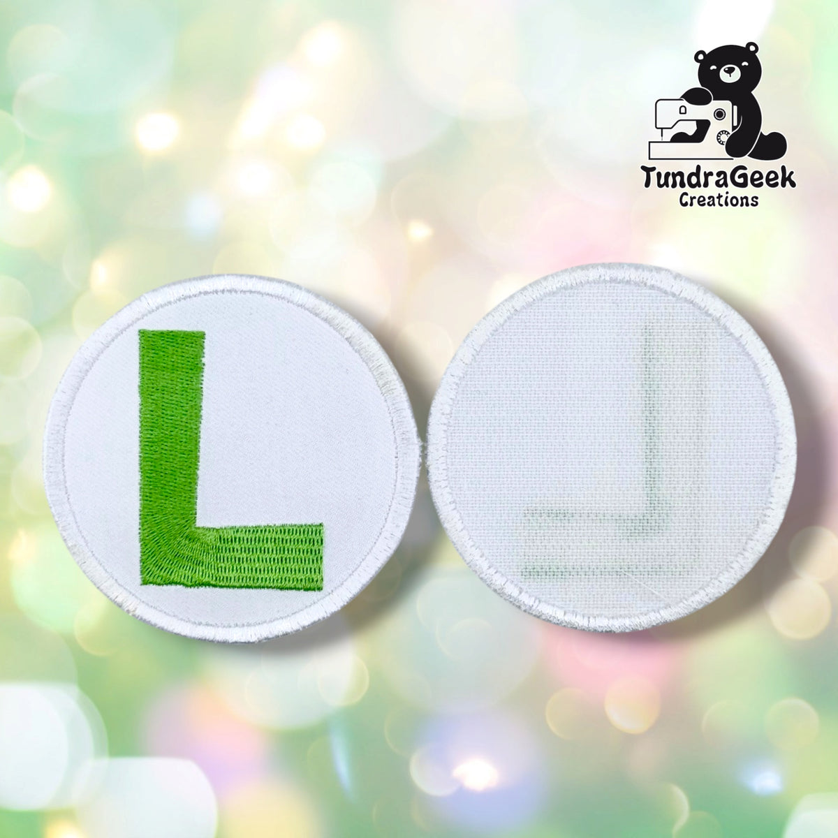 Two circular embroidered letter patches shown side by side on a soft pastel bokeh background. The left patch shows a green embroidered letter L on a white fabric base, and the right patch shows the reverse side with a light gray embroidered corner detail.
