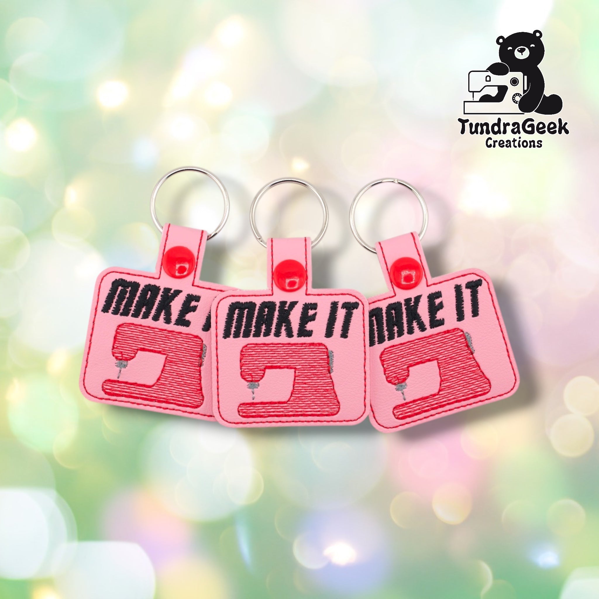 Three Make It Sew keychains arranged side by side against a soft bokeh background, all on pink vinyl with matching red embroidery and red plastic snap closures.