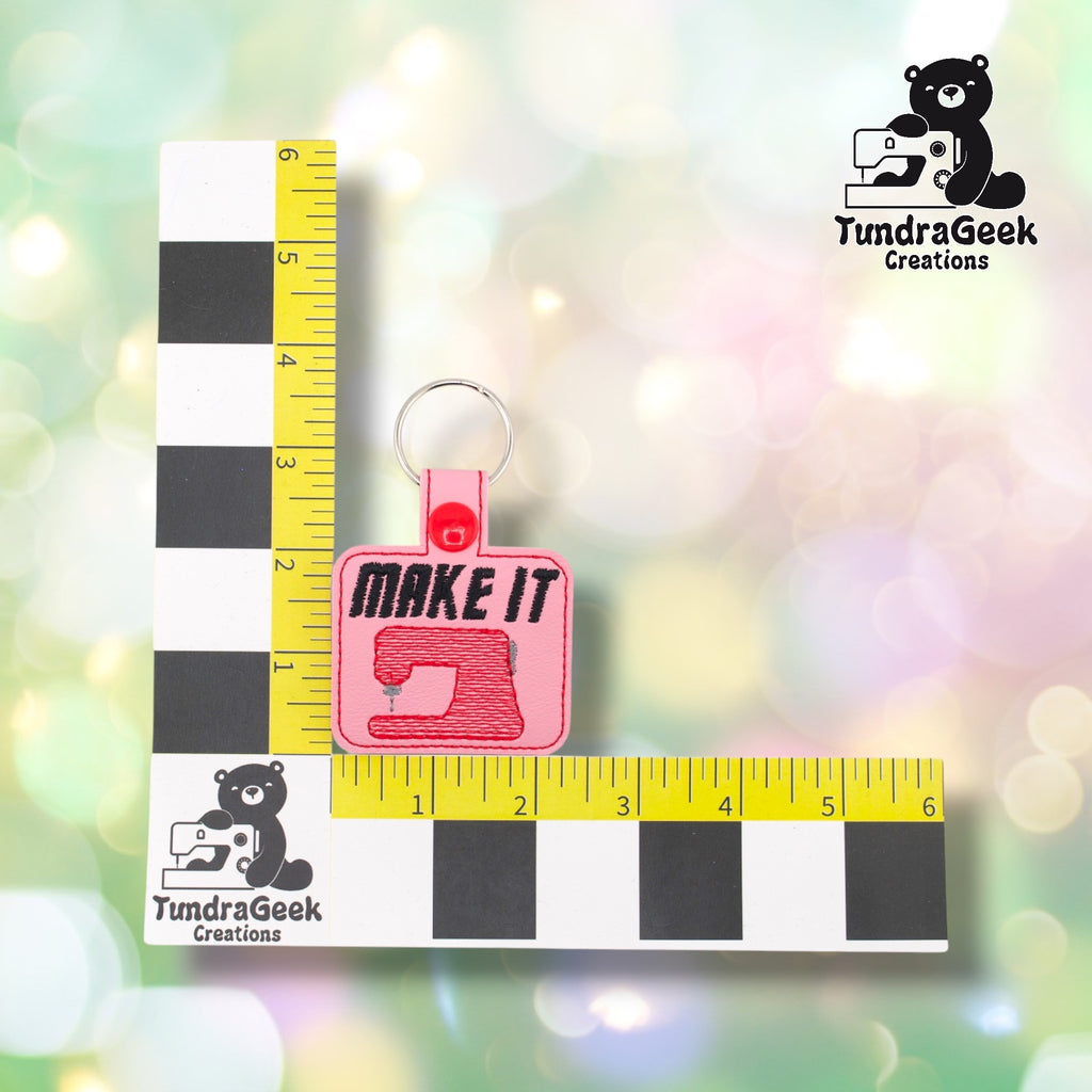 Pink Make It Sew keychain shown against a TundraGeek Creations L-shaped ruler prop. The keychain body measures just under 2.5 inches wide by 2 inches tall. Total height including tab and split ring is approximately 3.75 inches.