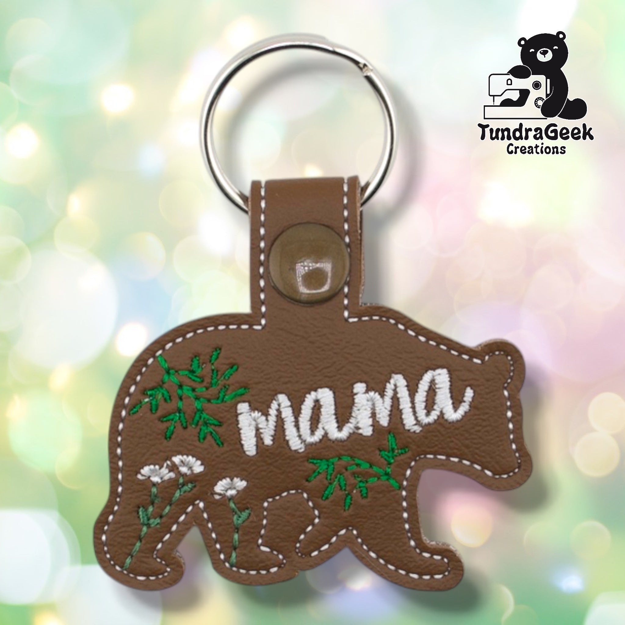 Brown faux leather keychain shaped like a walking bear. White lowercase script embroidery reads “mama” across the body; green botanical and wildflower embroidery fills the remaining space. Olive plastic snap closure and silver key ring at top. TundraGeek Creations logo in upper right corner.