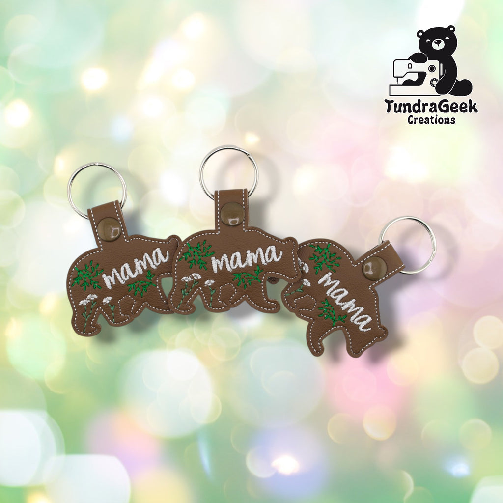 Three Mama Bear keychains arranged in a row on a pastel bokeh background, showing the front design from slightly varied angles. All three feature matching brown faux leather, white “mama” script, and green botanical embroidery.