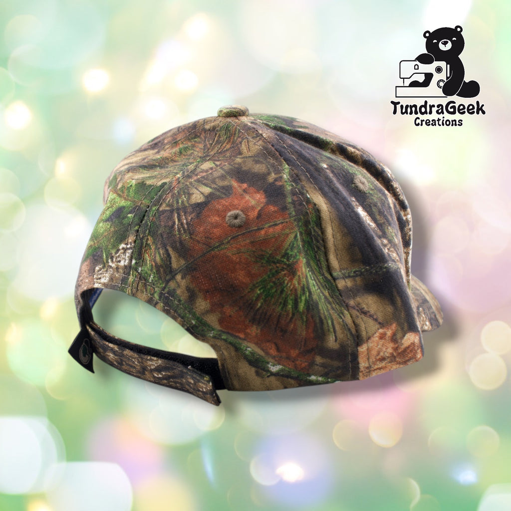 A camouflage baseball cap with brown, green, and tan forest pattern, displayed against a soft blurred pastel background with the TundraGeek Creations logo.