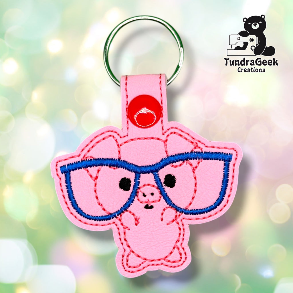 Front view of a pink vinyl keychain featuring an embroidered pig face wearing blue-rimmed glasses, with black stitched eyes, a pink stitched snout with nostrils, and pink stitched ear and body outlines, finished with a red plastic snap closure and silver split ring.