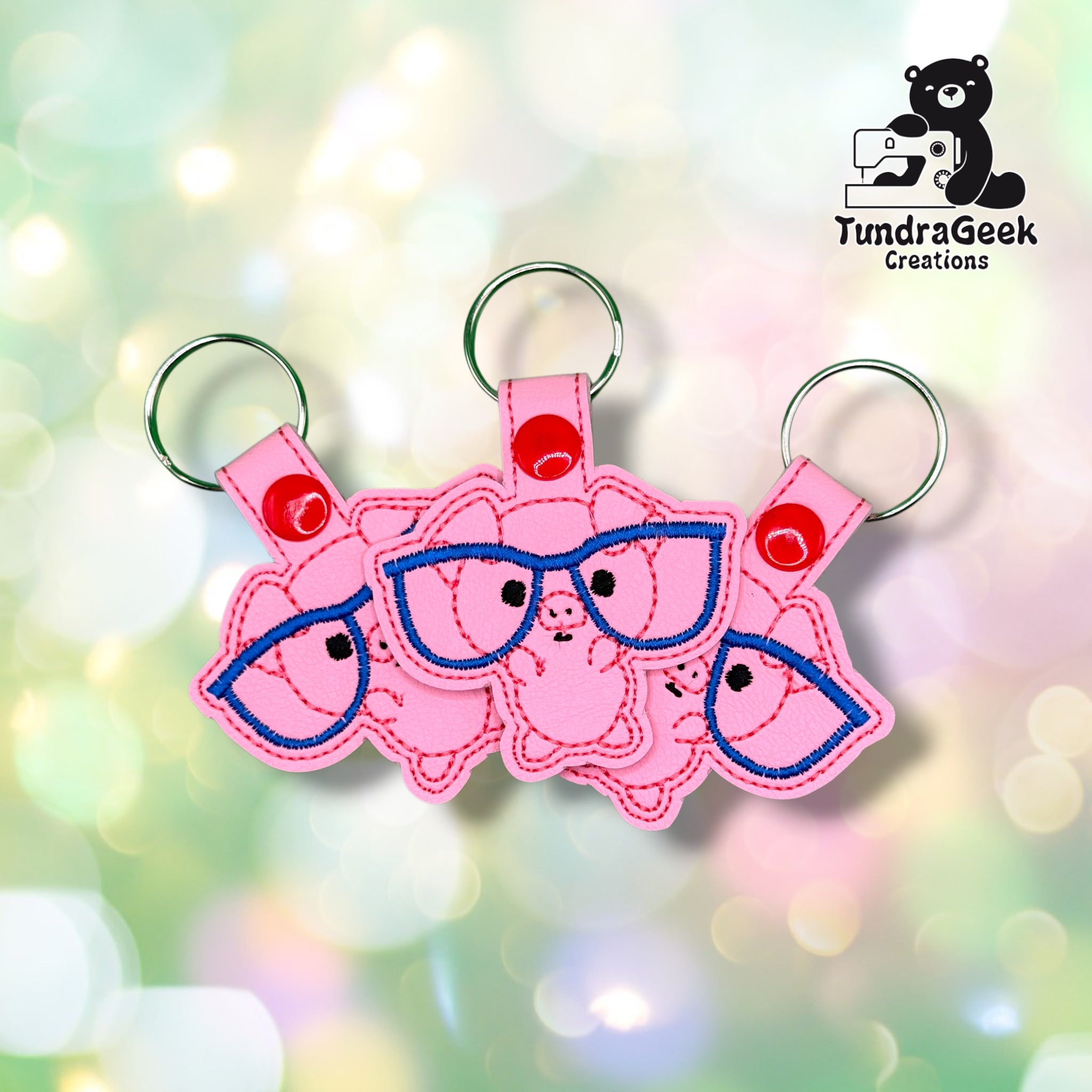 Three of the same keychain fanned out together, each showing the pig wearing blue-rimmed glasses on pink vinyl, with red plastic snap closures and silver split rings.