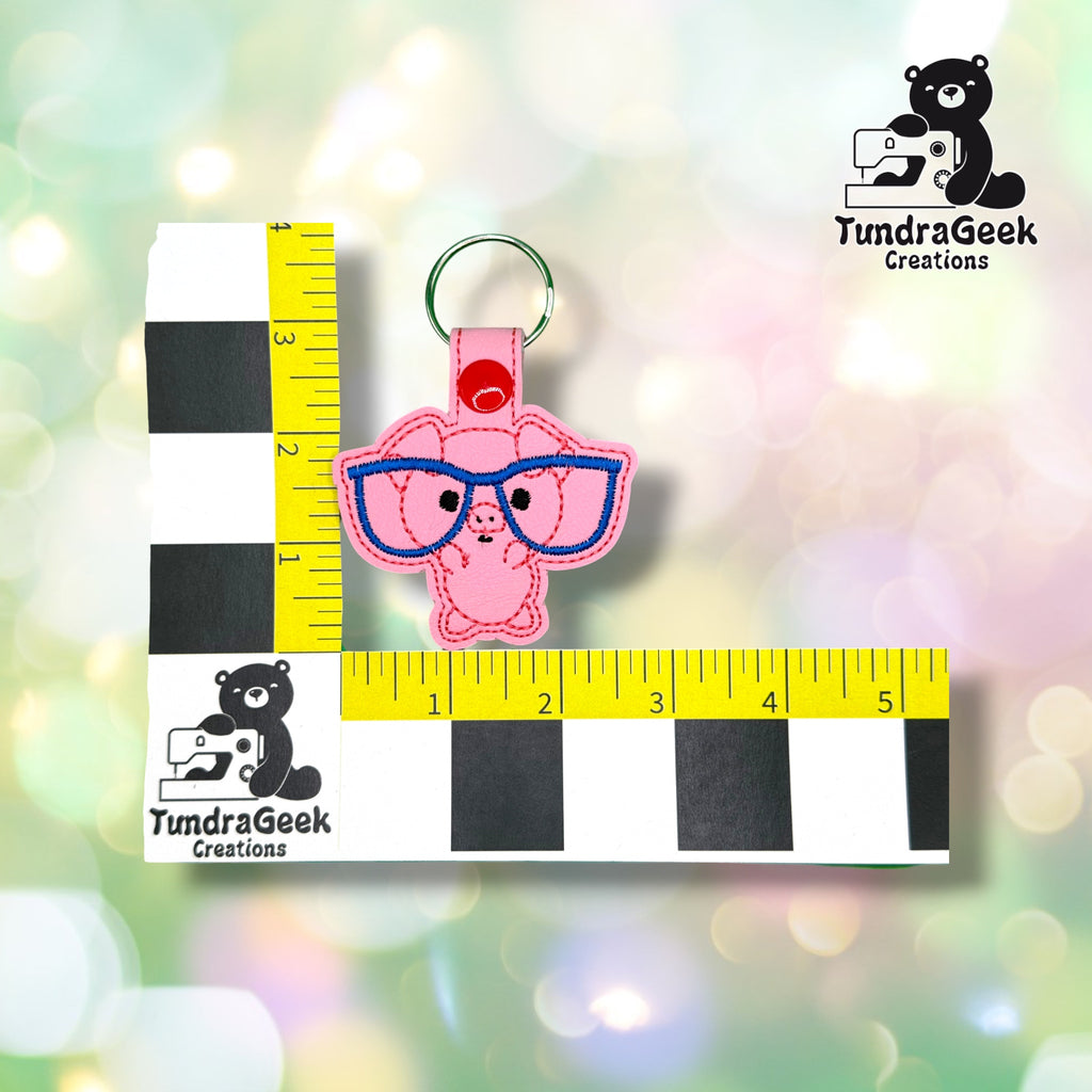 Keychain shown against a black-and-white checkered background with yellow rulers for scale, measuring approximately 4 inches tall by 3 inches wide including hardware.  ---