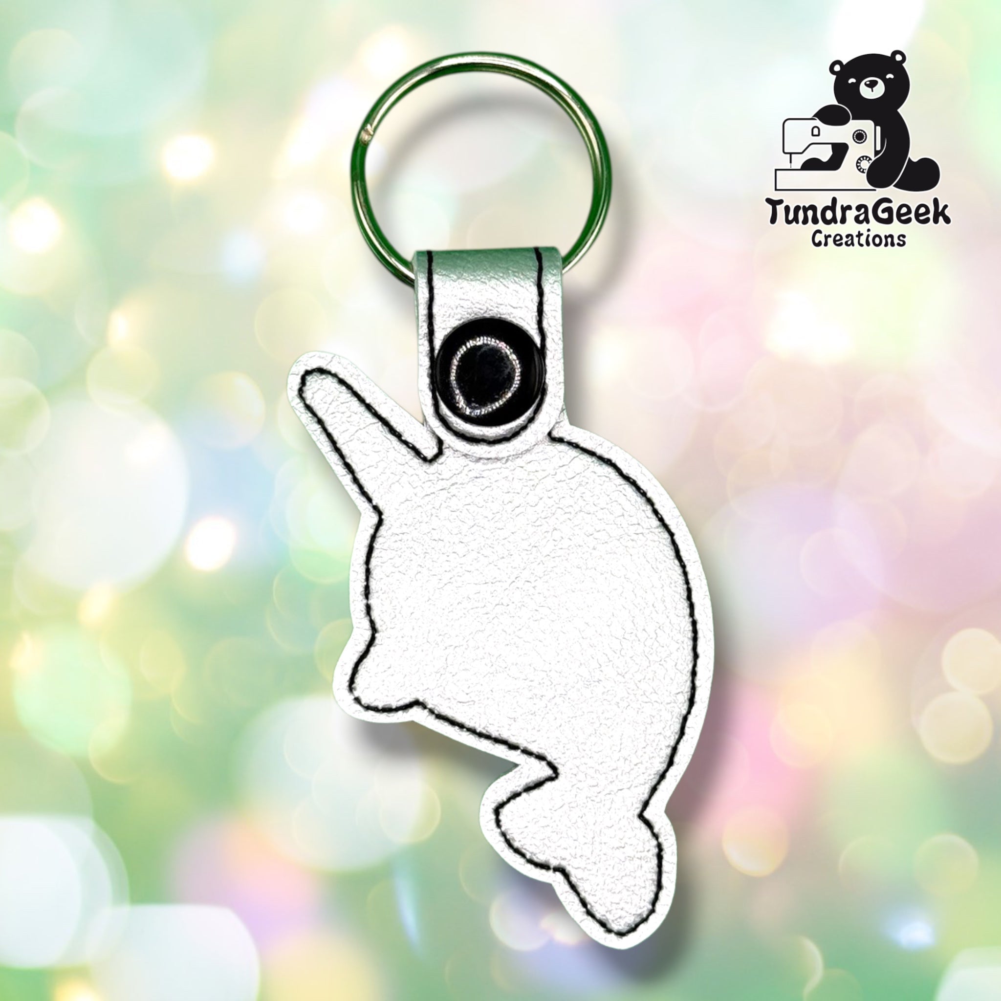 Back of a white vinyl keychain die-cut in the narwhal's body shape, showing a plain reverse side with black outline stitching following the silhouette. Green metal split ring and black snap closure at top.