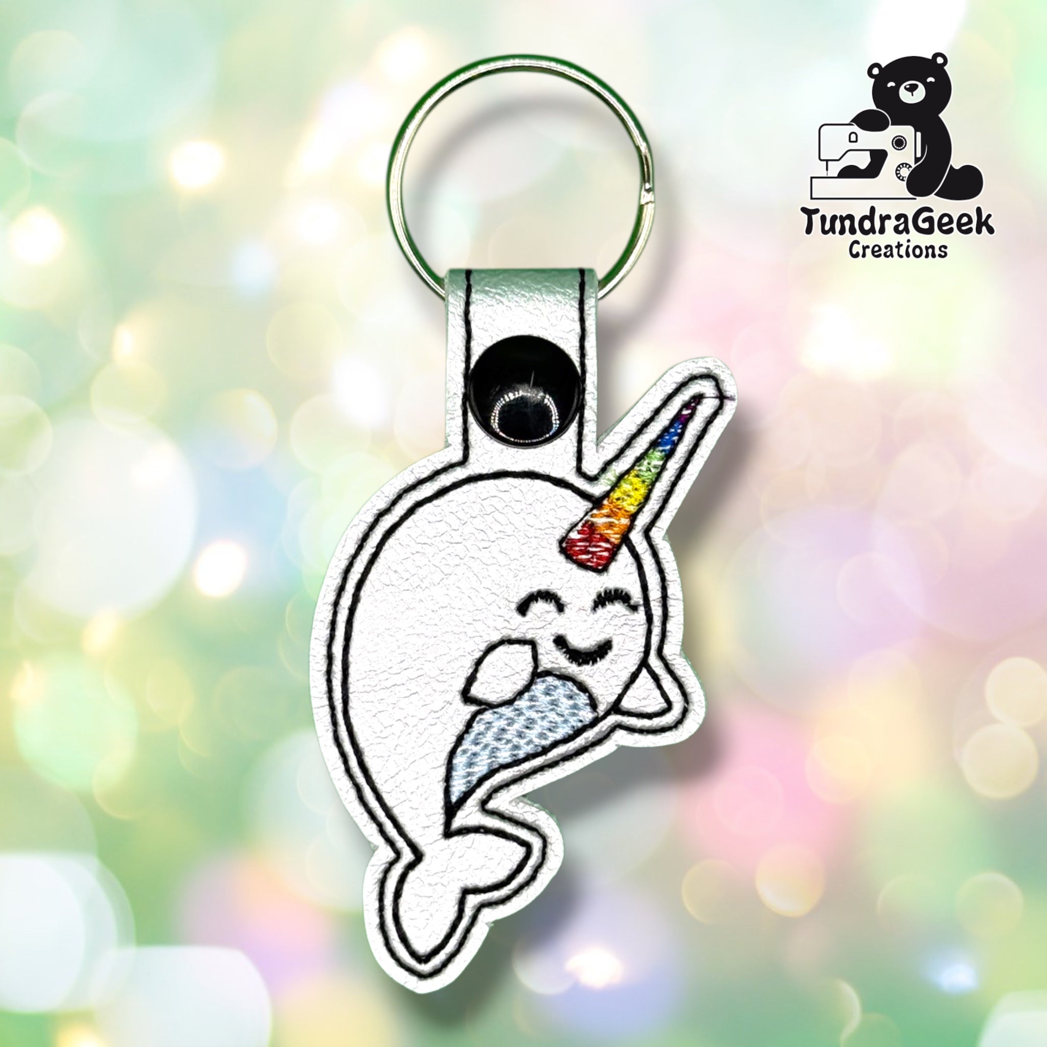 Front of a white vinyl keychain embroidered with a narwhal, featuring closed eyes and a content smile, a rainbow-striped horn, and a soft gray-and-white patterned tail. Green metal split ring and black snap closure at top.