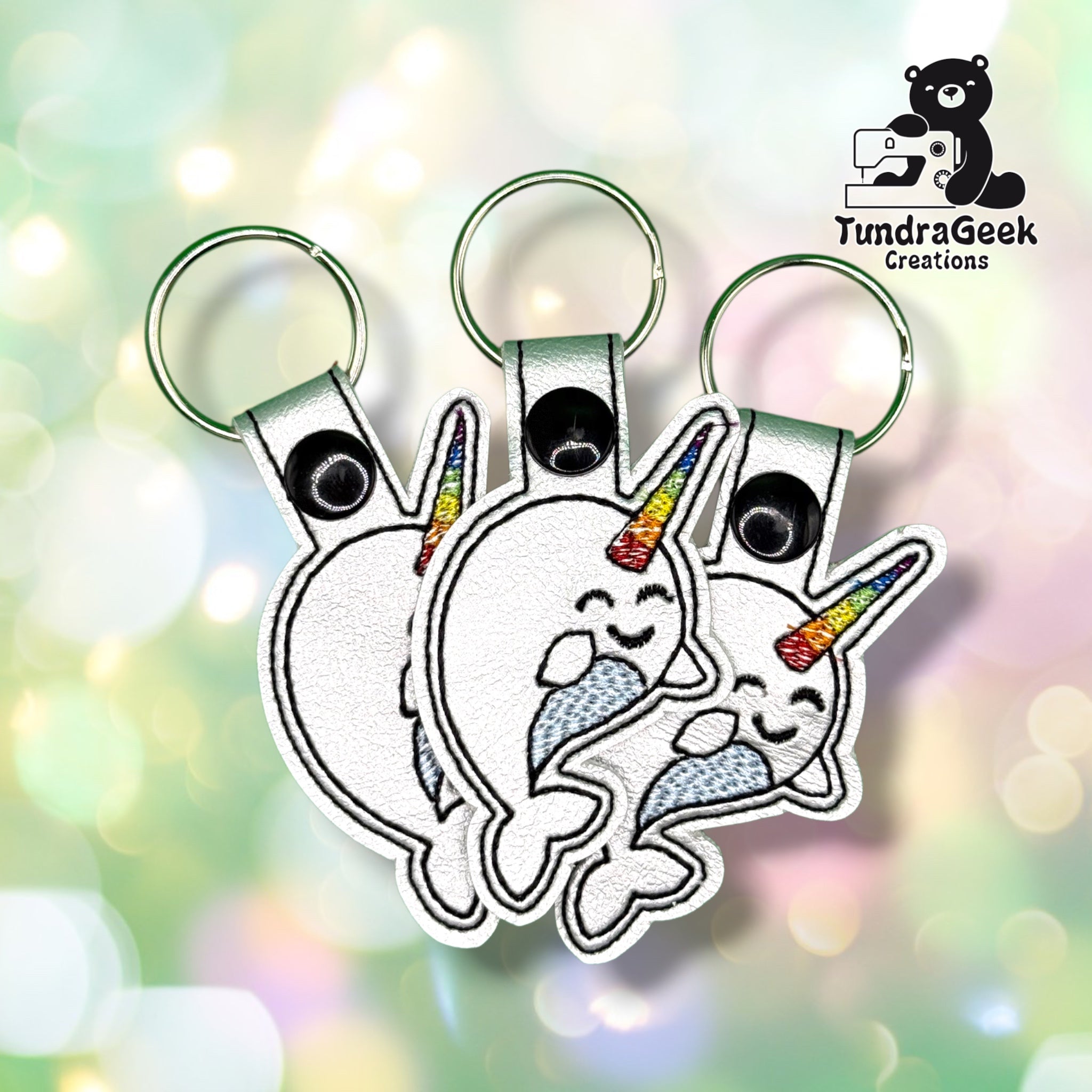 Three identical rainbow narwhal keychains fanned out together, each on a green metal split ring, showing the same white vinyl body, rainbow horn, and patterned tail.