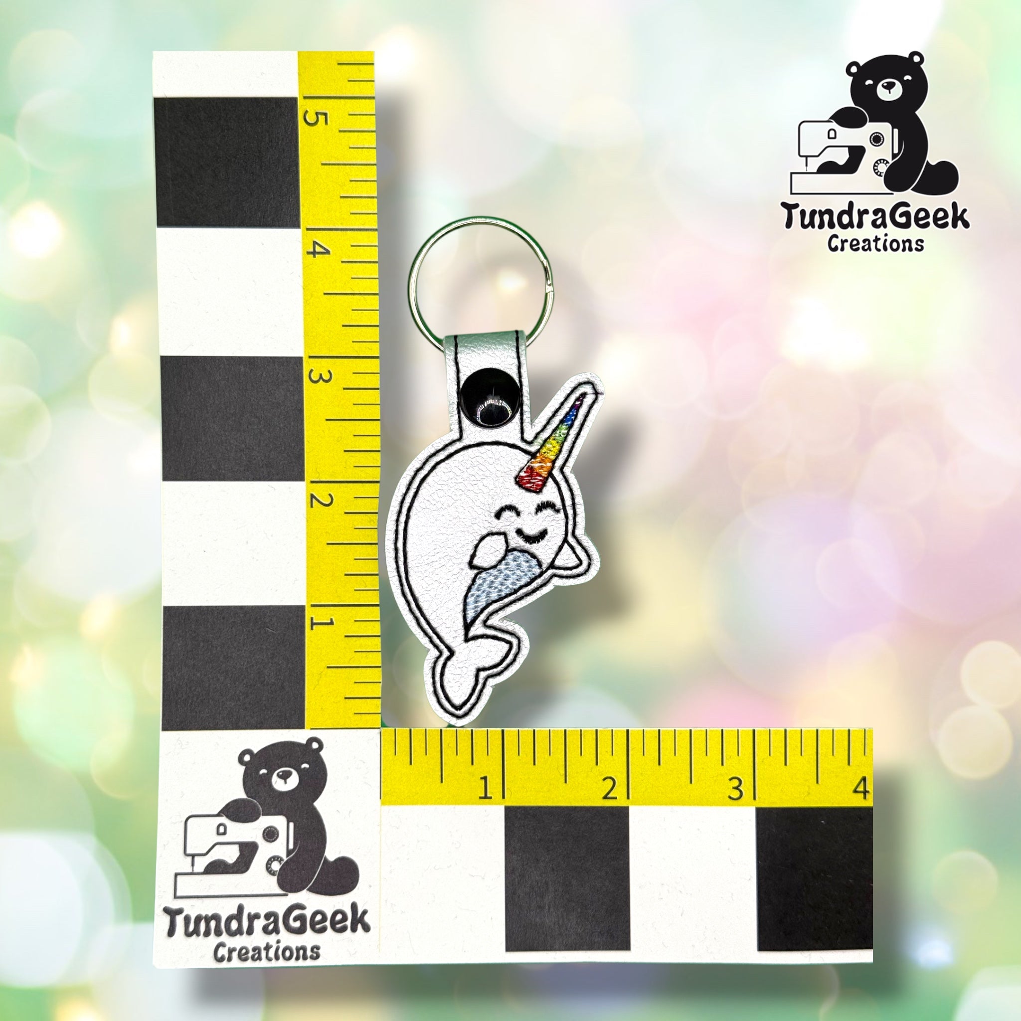 Rainbow narwhal keychain shown against a black-and-white checkered background with a yellow ruler for scale, measuring approximately 4.25 inches tall by 2 inches wide.  ---