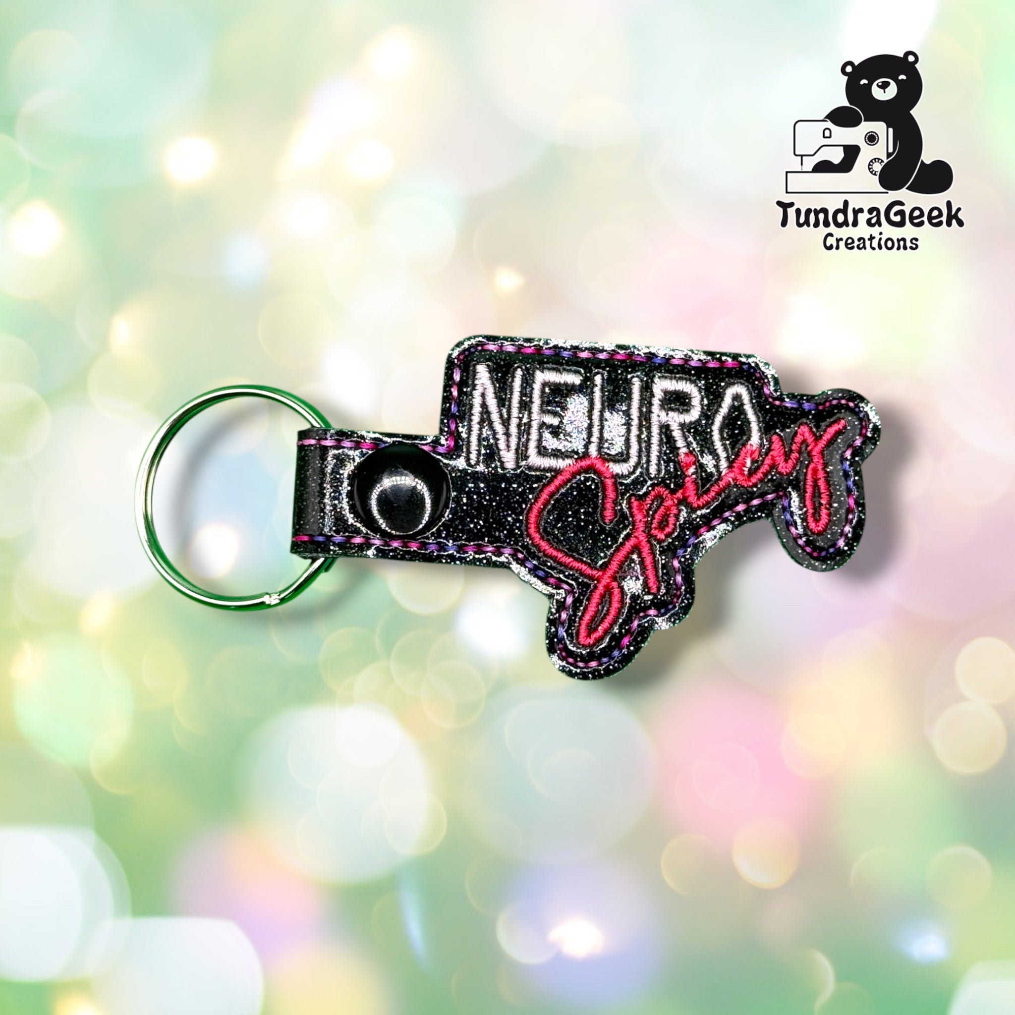 Front view of a black glitter vinyl keychain shaped to the design outline, with "NEURO" embroidered in pink block lettering and "Spicy" embroidered in pink script beneath it, finished with pink and purple stitched edge detailing and a black plastic snap closure.