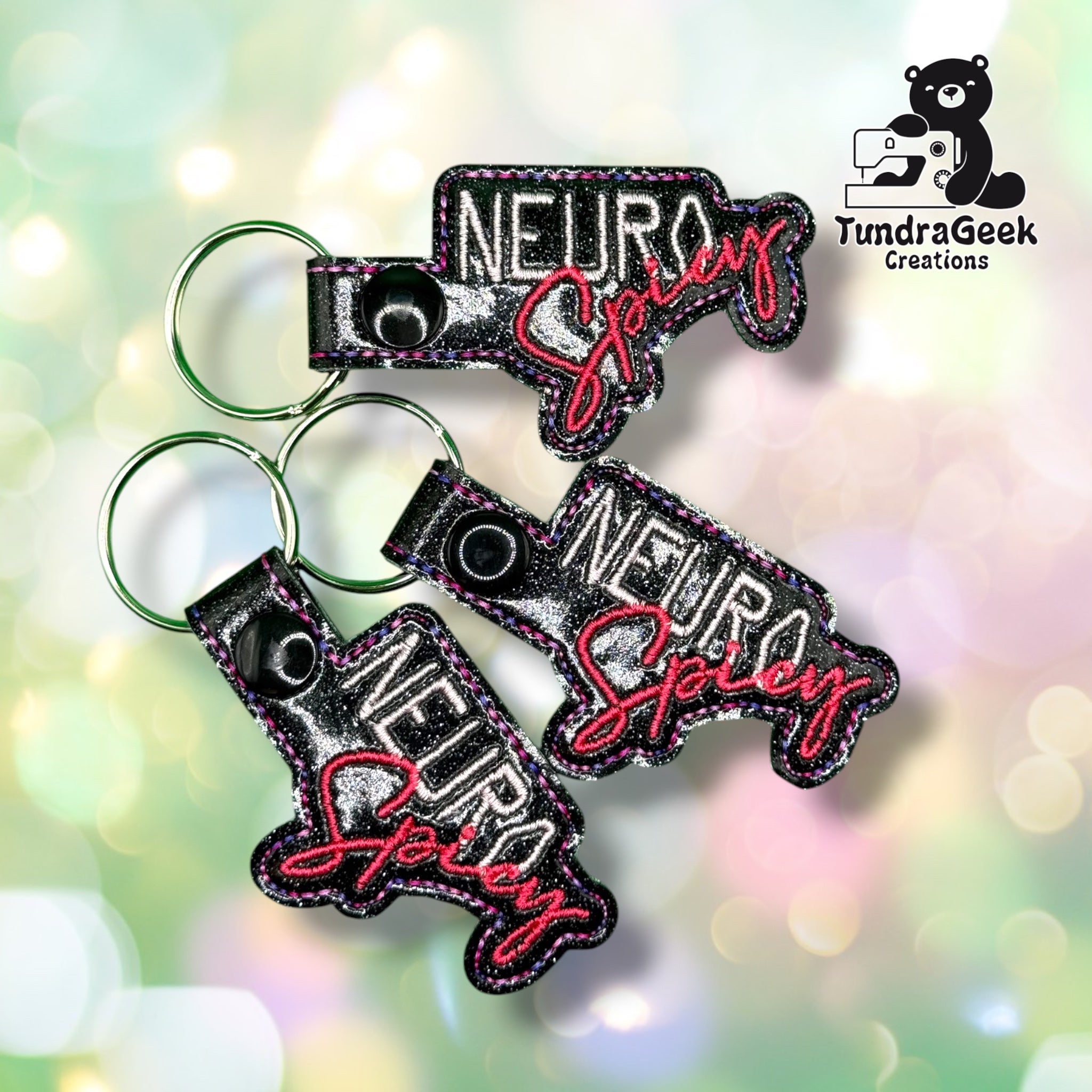 Three of the same keychain shown together, each black glitter vinyl with "NEURO" in white block letters and "Spicy" in pink script, pink and purple stitched edges, and black plastic snap closures.