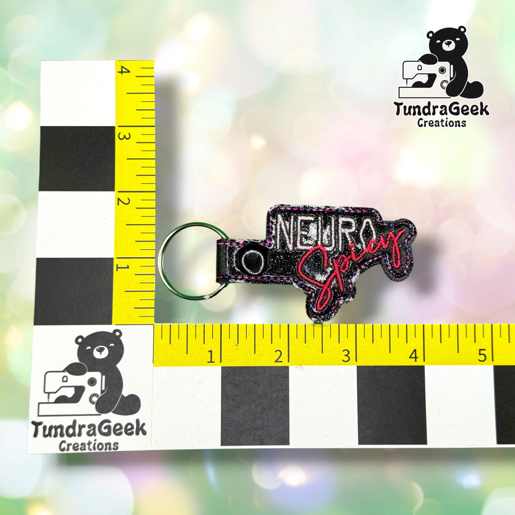 Keychain shown against a black-and-white checkered background with yellow rulers for scale, measuring approximately 4 inches wide by 2 inches tall including hardware.