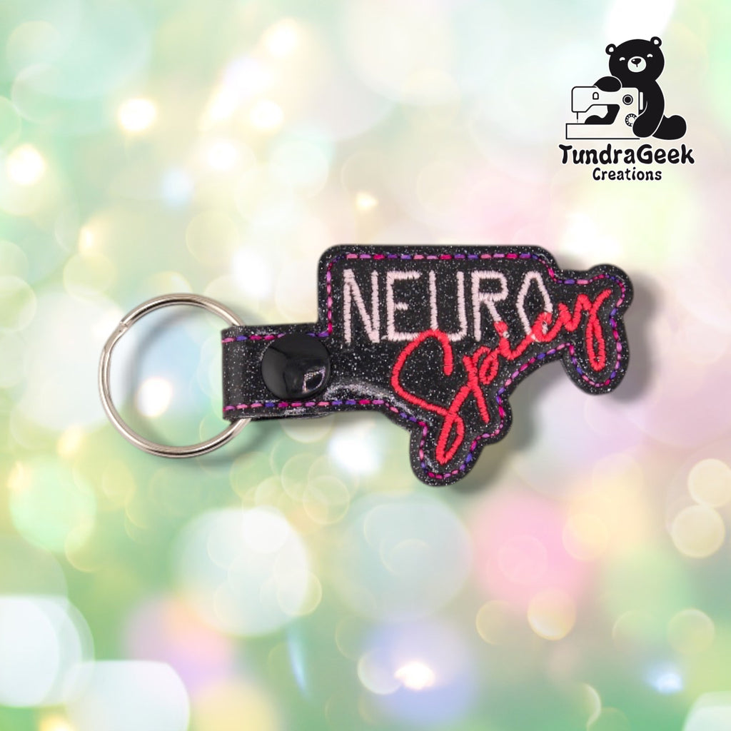 Front view of a black glitter vinyl keychain die-cut to the design outline, with "NEURO" embroidered in pale pink block lettering above "Spicy" in red-coral script, finished with pink and purple double-stitched edge detailing and a black plastic snap closure with silver split ring.